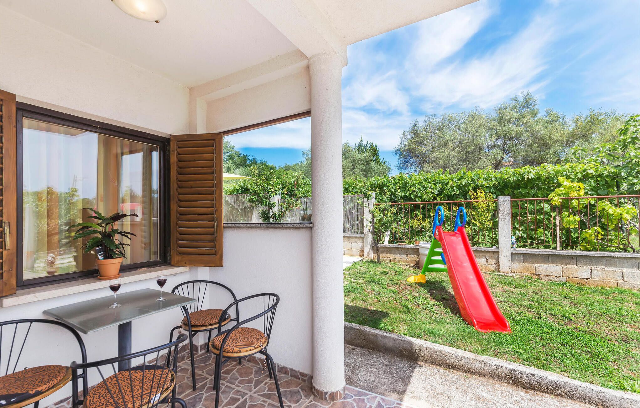 Un patio con mesa y sillas, una ventana con contraventanas y vistas a un jardín con un tobogán.