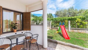 Um pátio com mesa e cadeiras, uma janela com portadas, e vista para um jardim com um escorrega.