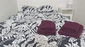 Una cama con una colcha floral en blanco y negro, dos toallas rojas oscuras dobladas y una mesita de noche blanca con una lámpara.