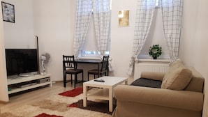 Una sala de estar con un sofá, un televisor, un escritorio con dos sillas y una ventana con cortinas.