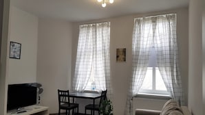 Una sala de estar con sofá, televisor, mesa de comedor con sillas y dos ventanas con cortinas.