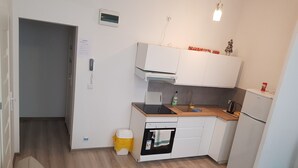 Una cocina compacta con armarios blancos, un lavamanos pequeño y un refrigerador. Hay un bote de basura amarillo y una mesita con una planta.