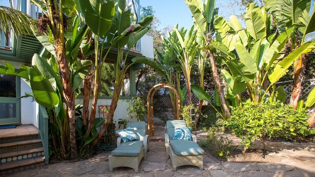 A garden with two wicker lounge chairs with cushions, surrounded by large banana plants and a wooden archway. - Santa Monica Beach Luxury Suite (Santa Monica)