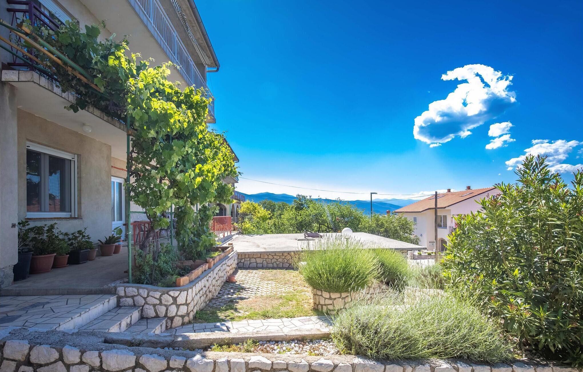 Une cour avec des marches en pierre menant à une maison dotée d’un balcon, entourée de plantes en pot et surplombée d’un ciel bleu clair.