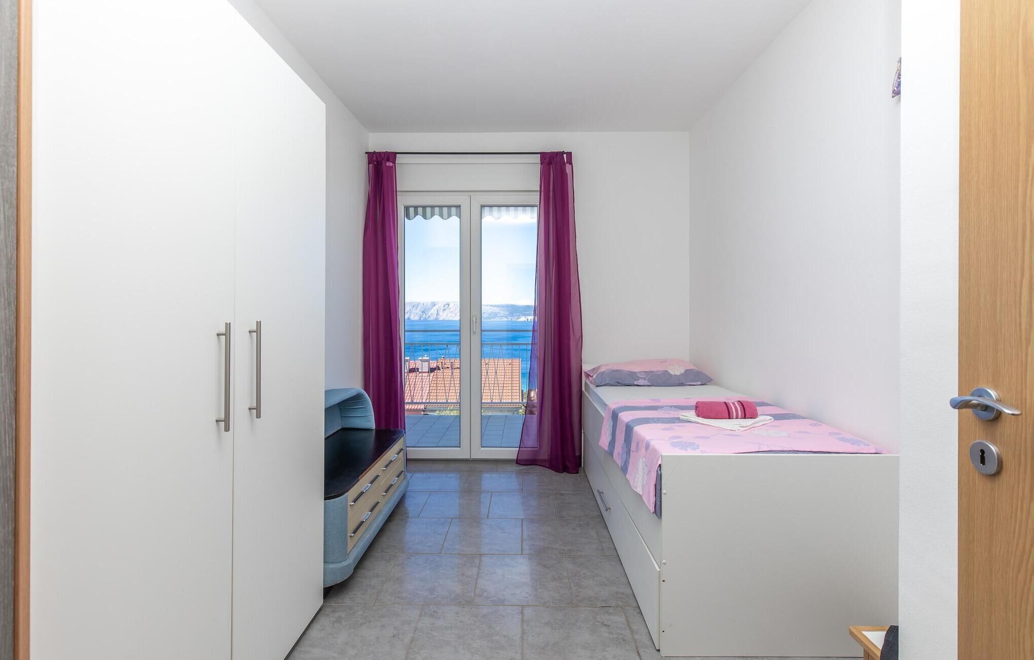 A small, bright room with a bed, a wardrobe, and a sliding glass door leading to a balcony with a view of the sea.