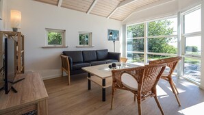 Ein modernes Wohnzimmer mit einem dunkelgrauen Sofa, einem Glastisch, Korbstühlen und großen Fenstern, die einen Blick ins Grüne bieten.