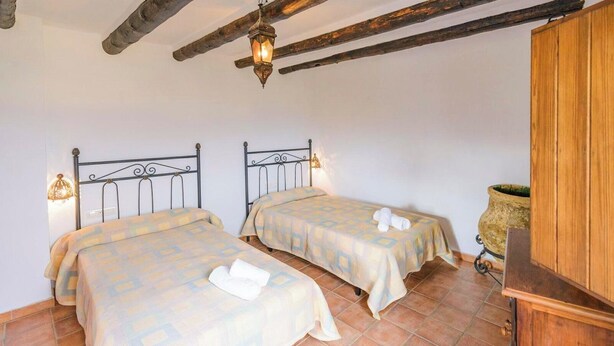 A room with two beds, each with a headboard and a bedside lamp. There is a wooden cabinet and a large potted plant in the corner. - Dehesa de las Casas Algarinejo by Ruralidays (Granada)