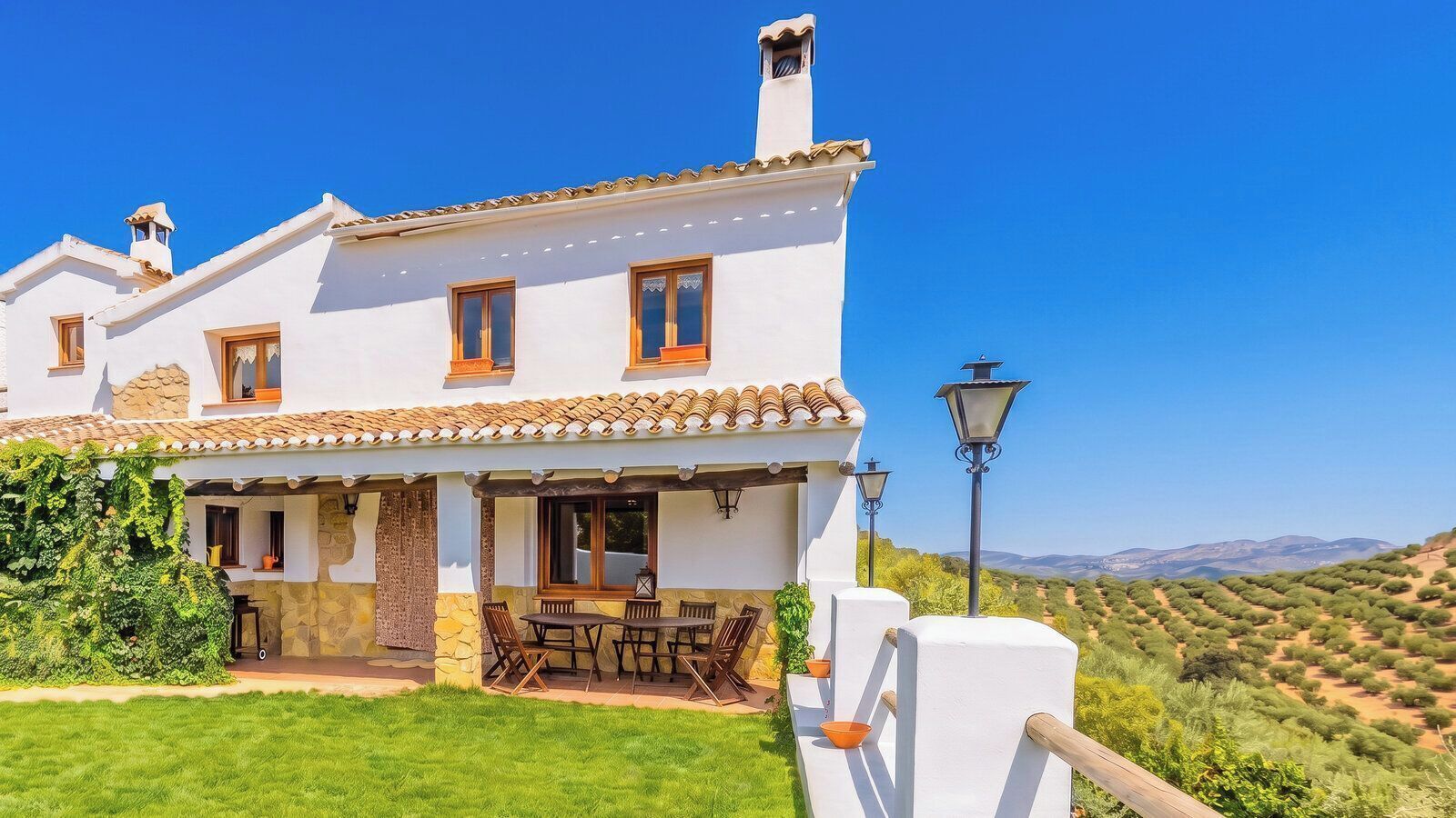 Una villa blanca con techo de tejas, un patio con mobiliario exterior y vistas a un paisaje montañoso.