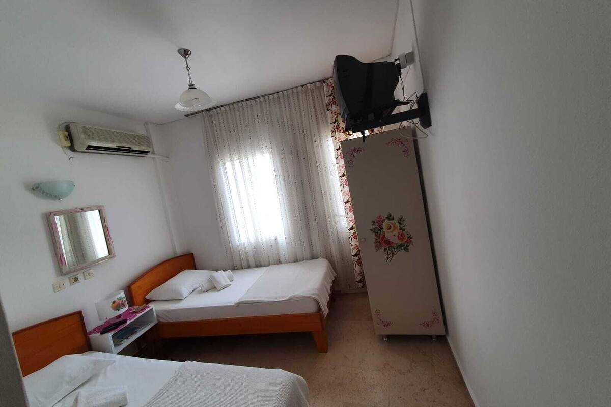 Quarto pequeno e limpo com duas camas individuais, guarda-roupa, espelho, ar condicionado e ventoinha de teto.