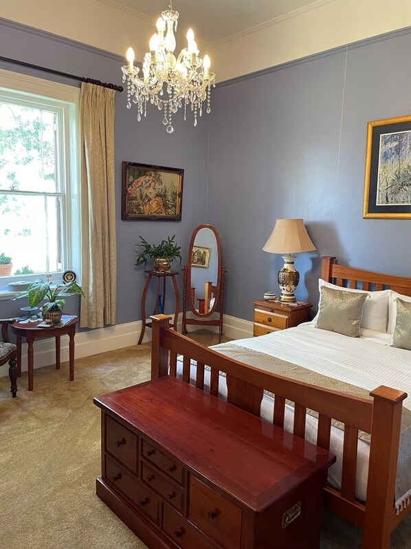 A bedroom with a wooden bed, a chandelier, a nightstand, a lamp, a dresser, a mirror, and a window with curtains.