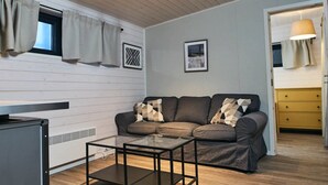 A modern living room with a glass coffee table, a sofa with patterned cushions, and a kitchen area visible through an open door.