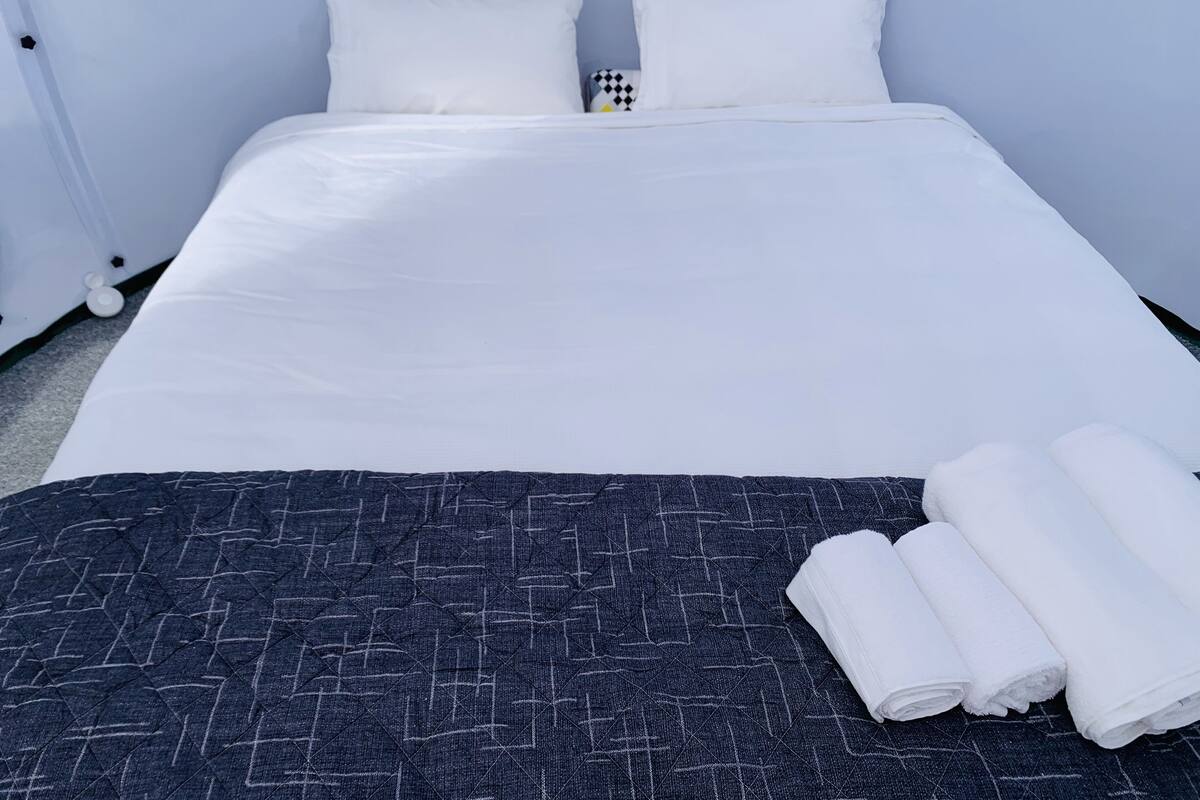 Een netjes opgemaakt bed met witte lakens, een donkerblauwe quilt en twee opgerolde witte handdoeken.