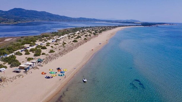 Vista aérea de uma praia com águas azul-claras, faixa arenosa e fileira de cabanas à beira-mar.