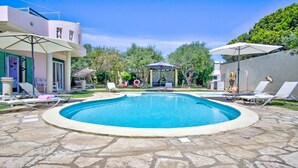 Uma piscina com espreguiçadeiras e guarda-sóis, rodeada por um pátio de pedra e um edifício com design moderno.