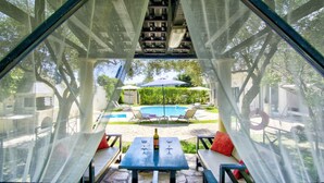 Vista pela janela de uma área da piscina com mesa, cadeiras e uma garrafa de vinho.