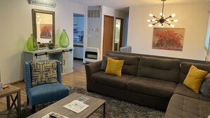 A living room with a brown sofa, blue armchair, and a coffee table with magazines and remote controls. A kitchen is visible in the background. - Beautiful Bi-level. Game room with pool table, 5 UHDTVs, WiFi. Central heat/air. (Bushkill)