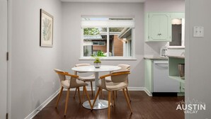 Un coin repas avec une table ronde et quatre chaises, une fenêtre donnant sur une maison, et des meubles de cuisine de couleur vert clair.