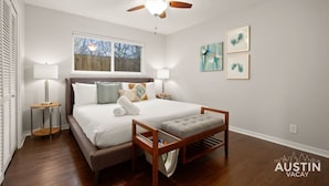 Une chambre à coucher comprenant un lit, des tables de chevet, un ventilateur de plafond, une fenêtre et des œuvres d’art accrochées au mur.