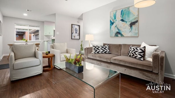 Un salon moderne avec une table basse en verre, un canapé beige agrémenté de coussins à motifs, un fauteuil blanc et une peinture abstraite contemporaine accrochée au mur.
