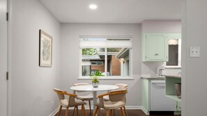 A dining area with a round table and four chairs, a window with a view of a house, and light green cabinets in the kitchen area.