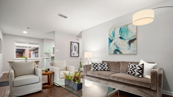 A modern living room with a glass coffee table, a beige sofa with patterned cushions, a white armchair, and a contemporary abstract painting on the wall.