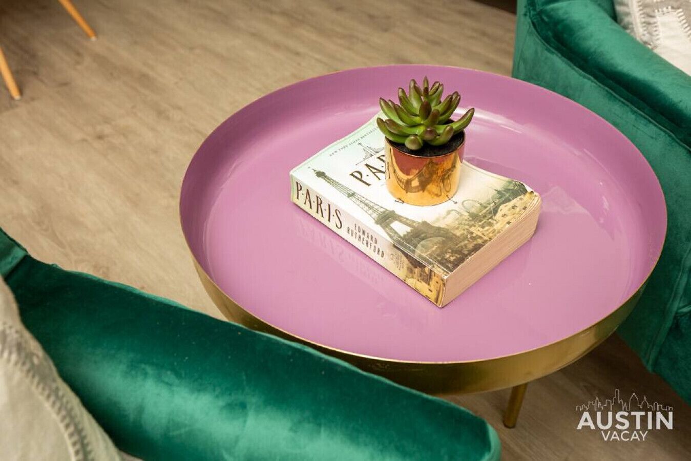 Een kleine ronde tafel met een paars blad, een gouden voet en een groene kussentje. Op de tafel ligt een boek met de titel 'PARIS' en een kleine plant in een pot.