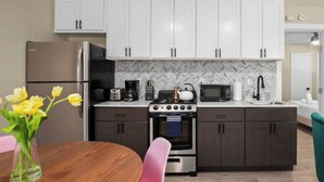 A modern kitchen with white cabinets, a stainless steel refrigerator, a gas stove, and a microwave. There is a wooden dining table with a vase of flowers. - 7-Minute Walk to Deep Eddy Pool! Parking + Grill (Austin)