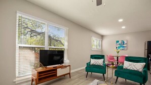 A living room with a TV on a wooden stand, two green armchairs, a small table with a vase, and a painting on the wall. - 7-Minute Walk to Deep Eddy Pool! Parking + Grill (Austin)