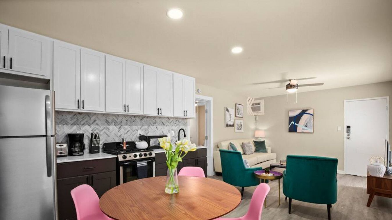 Une cuisine compacte avec des meubles blancs, un réfrigérateur en acier inoxydable et une cuisinière à gaz. On y trouve un coin repas avec une table en bois et des chaises roses. Le salon est doté d’un canapé turquoise et d’un fauteuil vert.