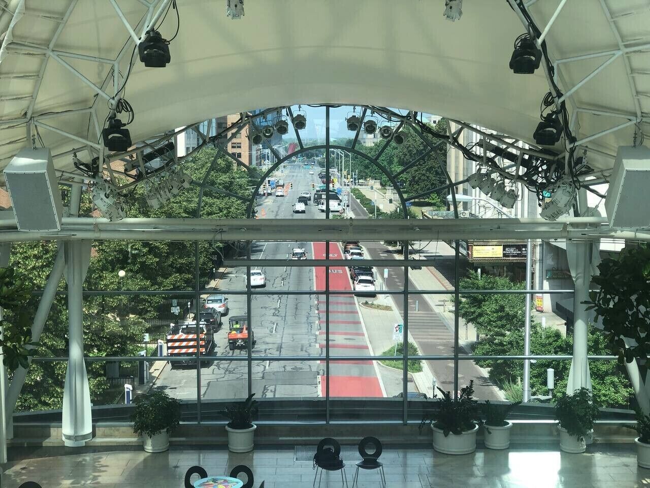 Vista a través de un cerramiento de vidrio con estructura metálica, plantas en macetas y una carretera con vehículos y un carril para autobuses rojo.