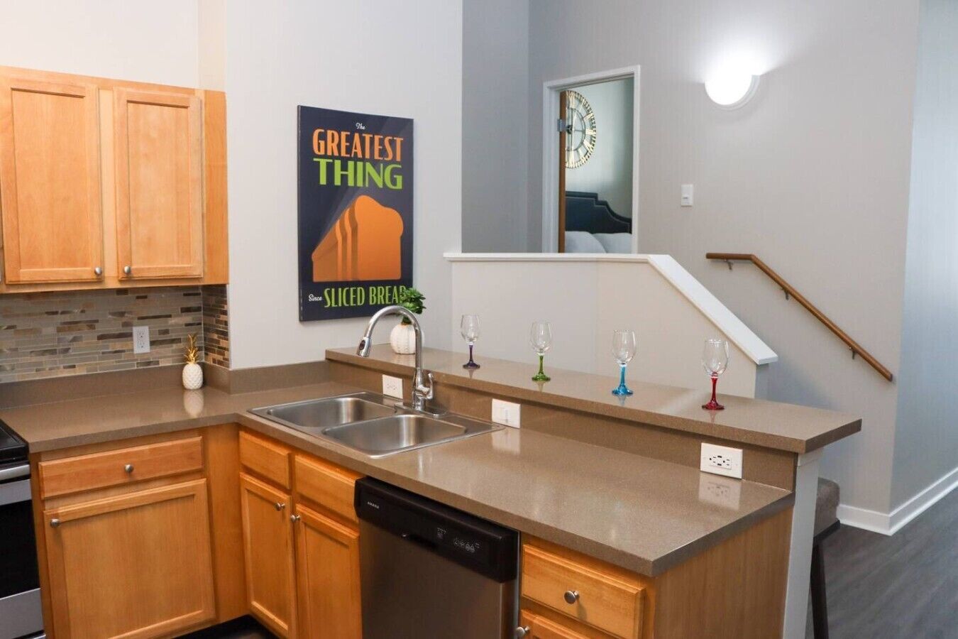 A modern kitchen with wooden cabinets, a dishwasher, a granite countertop, a poster on the wall, and a staircase with a clock.