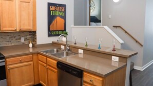 A modern kitchen with wooden cabinets, a dishwasher, a granite countertop, a poster on the wall, and a staircase with a clock. - EXECUTIVE STAY+KING BED+WIFI+W/D+PETS (Indianapolis)