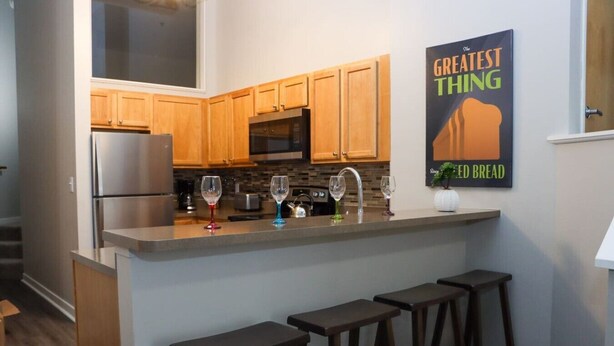 A modern kitchen with wooden cabinets, a stainless steel refrigerator, a microwave, and a bar area with stools. - EXECUTIVE STAY+KING BED+WIFI+W/D+PETS (Indianapolis)