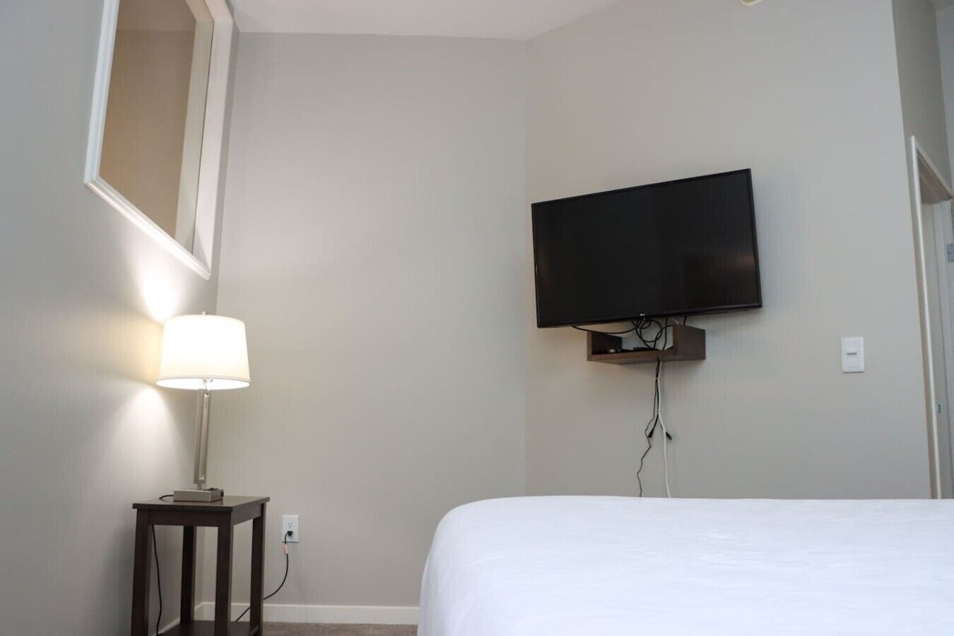 A hotel room with a bed, a wall-mounted TV, and a bedside table with a lamp.