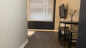 A hallway with a dark wood floor, a large window with blinds, a flat-screen TV on a stand, and a small table with chairs. - EXECUTIVE STAY+KING BED+WIFI+W/D+PETS (Indianapolis)