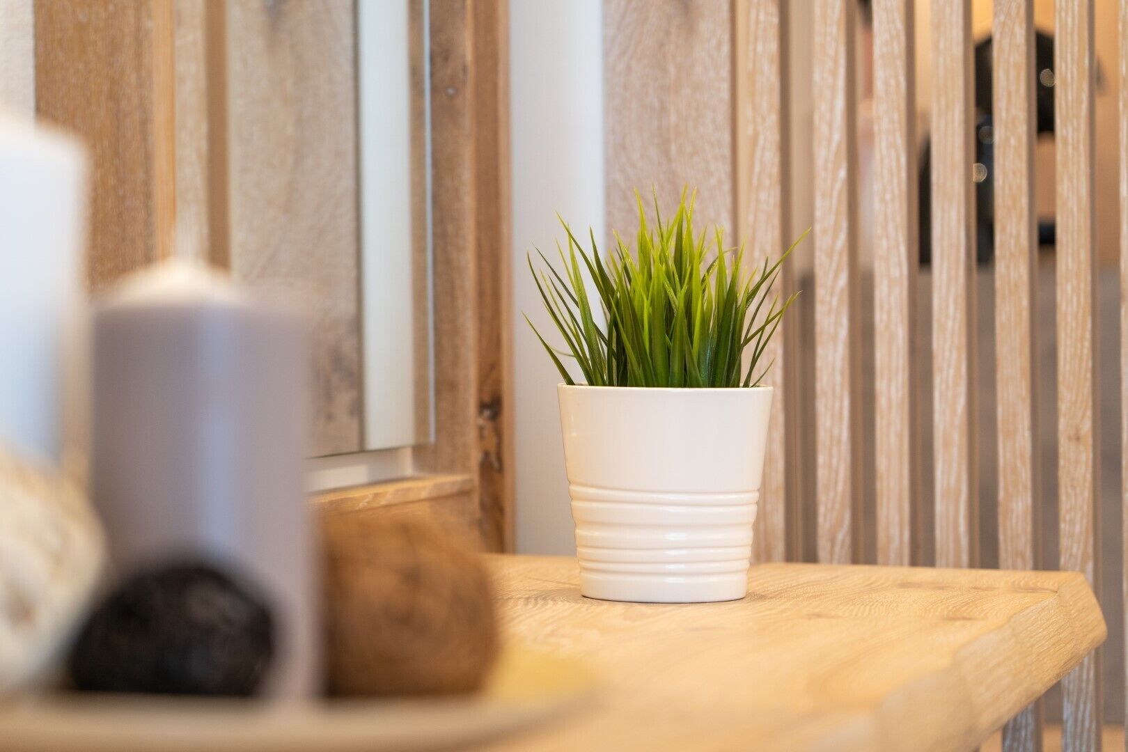 A wooden table with a white pot containing green plants, a white candle, and a dark object.