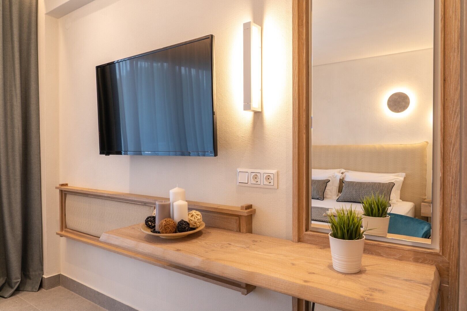 Un baño moderno con tocador de madera, televisor montado y un espejo que refleja una cama con detalles azules.