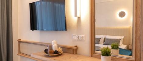 Un baño moderno con tocador de madera, televisor montado y un espejo que refleja una cama con detalles azules.