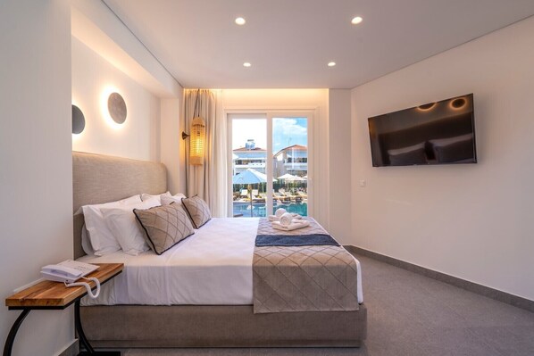 Habitación de hotel con cama, mesita de noche con teléfono, televisor montado en la pared y vista a una piscina y edificios.