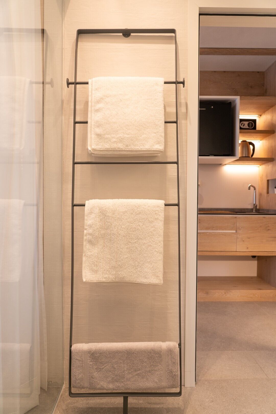 Un bagno con un portasciugamani con tre asciugamani, una cabina doccia in vetro e un mobile bagno in legno con lavandino e bollitore.