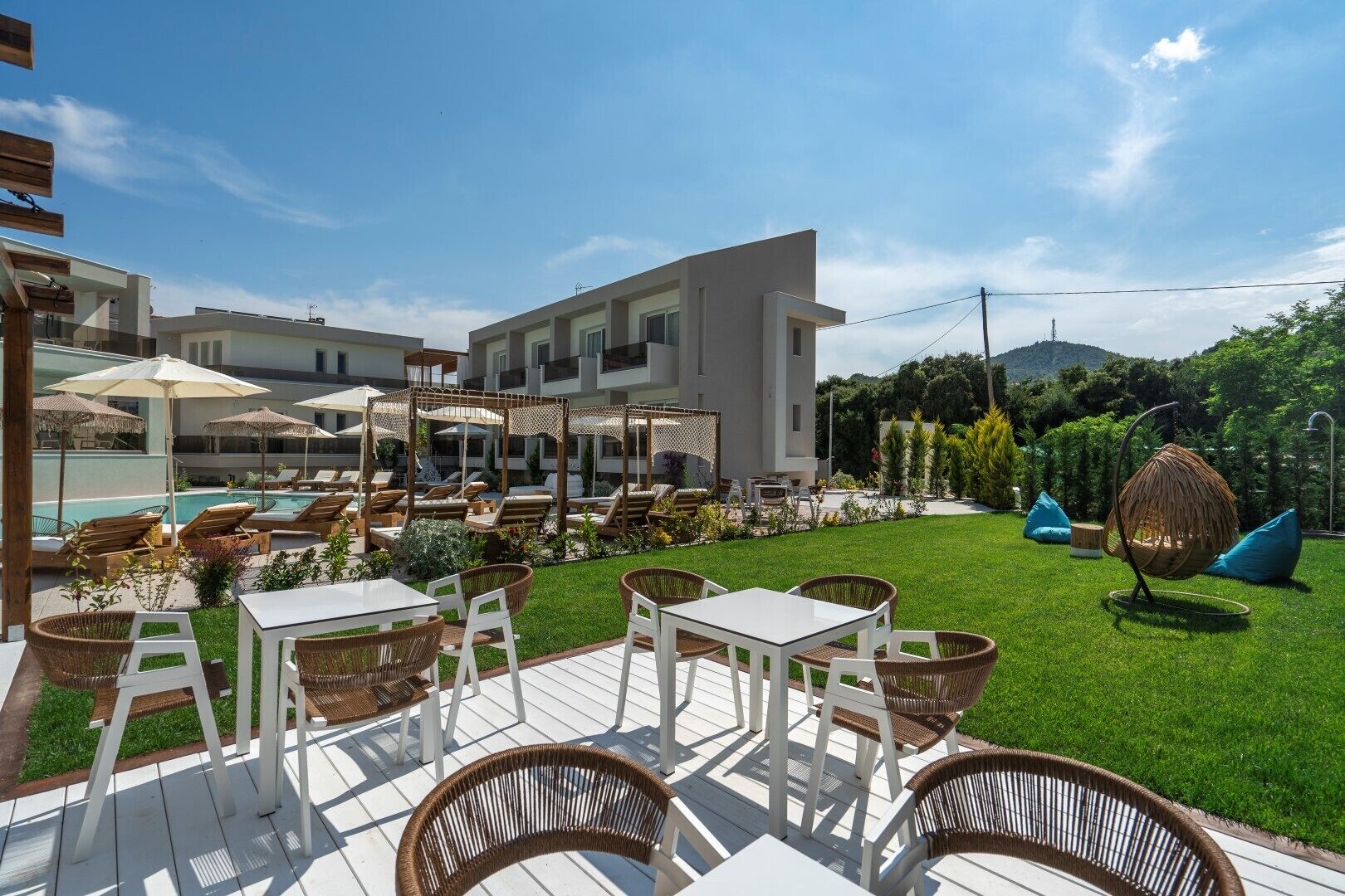 A modern outdoor patio with white tables and wicker chairs, a pool, and a well-maintained lawn.