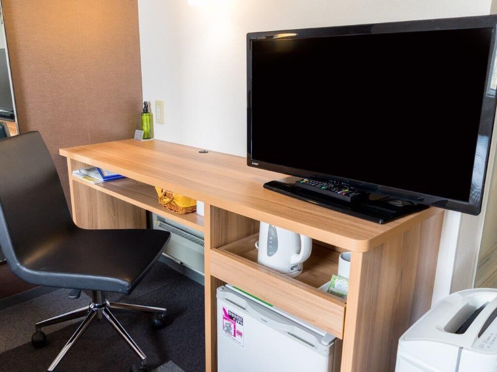 Un espace de travail dans une chambre d’hôtel, comprenant un bureau en bois, un fauteuil de bureau noir, un téléviseur à écran plat, un clavier, une machine à café et un mini-réfrigérateur.