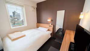 A hotel room with a bed, a desk with a TV, a mirror, and a window with a view of buildings.