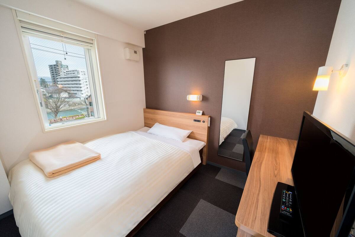 Habitación de hotel con cama, escritorio con televisor, espejo y ventana con vista a edificios.