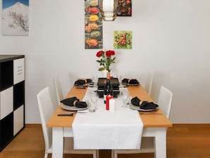 A dining area with a wooden table set for six, white chairs, a vase with red roses, and a wine bottle.