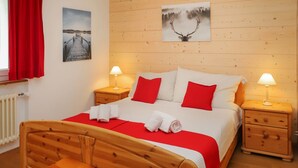 Een houten bed met rode beddengoed, twee nachtkastjes met lampen, een houten bankje en een schilderij van een hert op de muur.