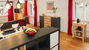 Een moderne woonkamer met een zwarte bank, een eettafel met een schaal fruit, een koffieapparaat en een boekenkast.