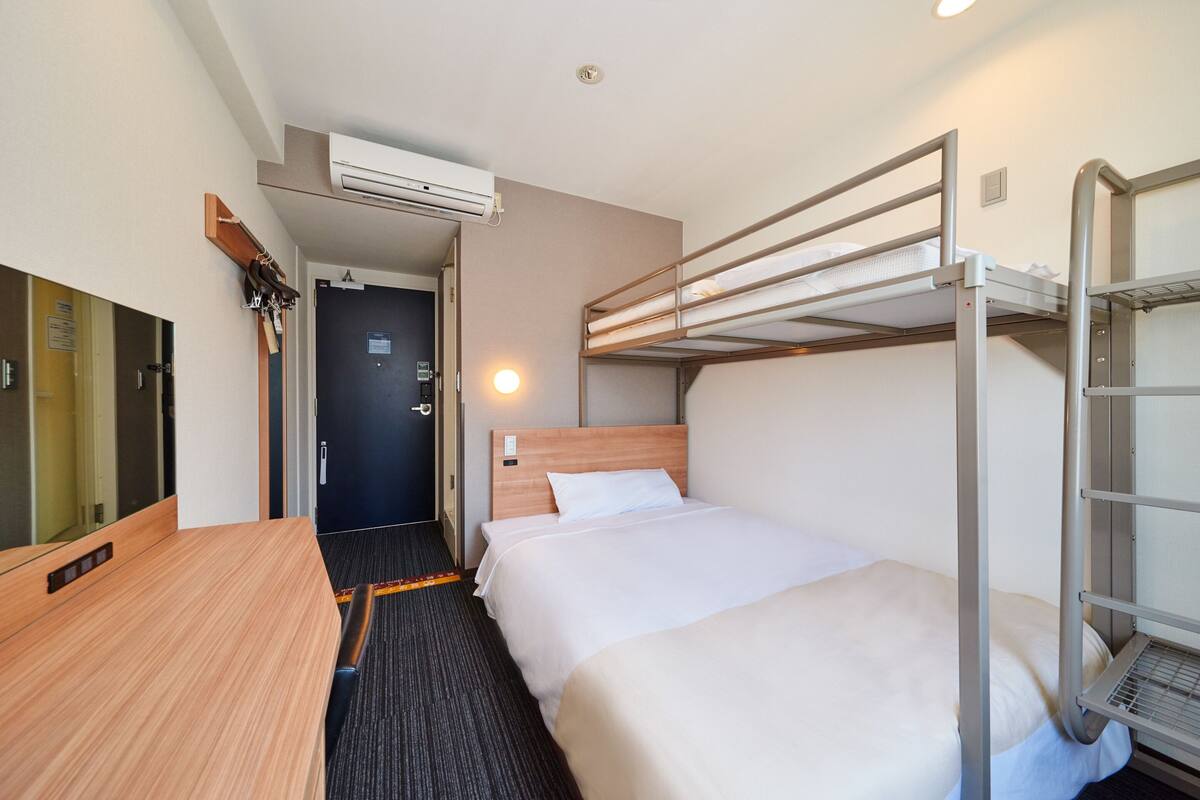 A hotel room with a bunk bed, a desk, a mirror, and an air conditioning unit.