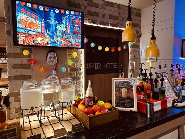 A bar area with a television displaying a cityscape, a sign reading 'SUPER IOTE,' and a variety of beverages and fruit.