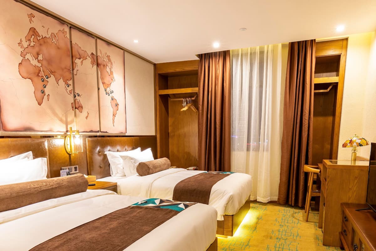 Une chambre d’hôtel avec deux lits, une carte du monde accrochée au mur, une tête de lit en bois, un bureau avec une lampe et une moquette à motifs.
